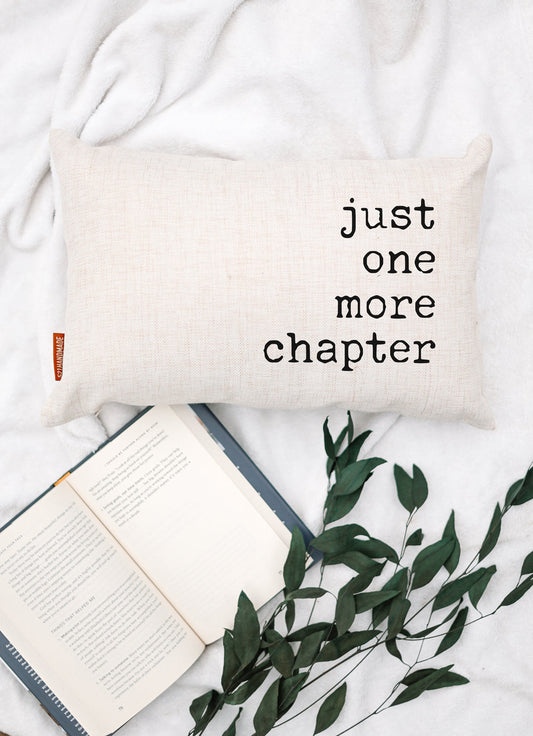 Cream fabric pillow with “just one more chapter” text beside an open book and green leaves